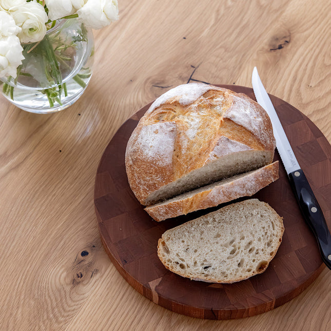 Loaf of bread sliced on an end-grain wooden cutting board with a knife, on a light wood countertop table with white flowers in the background.