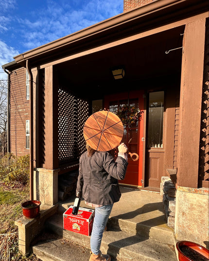 Person with a pizza cutting board and a bottle of wine standing on a porch with a blue sky background